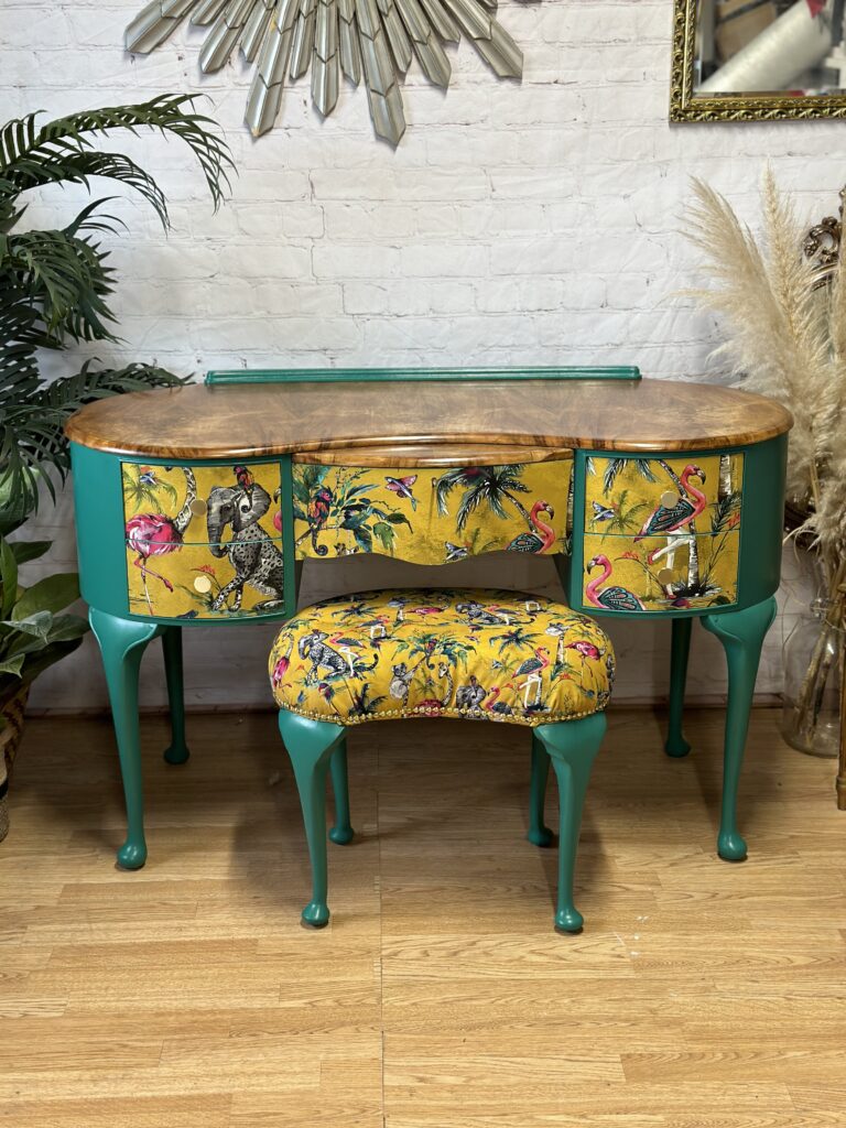 A vintage vanity table with vibrant green legs and sides, adorned with drawer fronts and a matching stool covered in yellow fabric with tropical bird and foliage patterns. The tabletop has a natural wood finish. Decorative elements include plants and a stylish mirror.