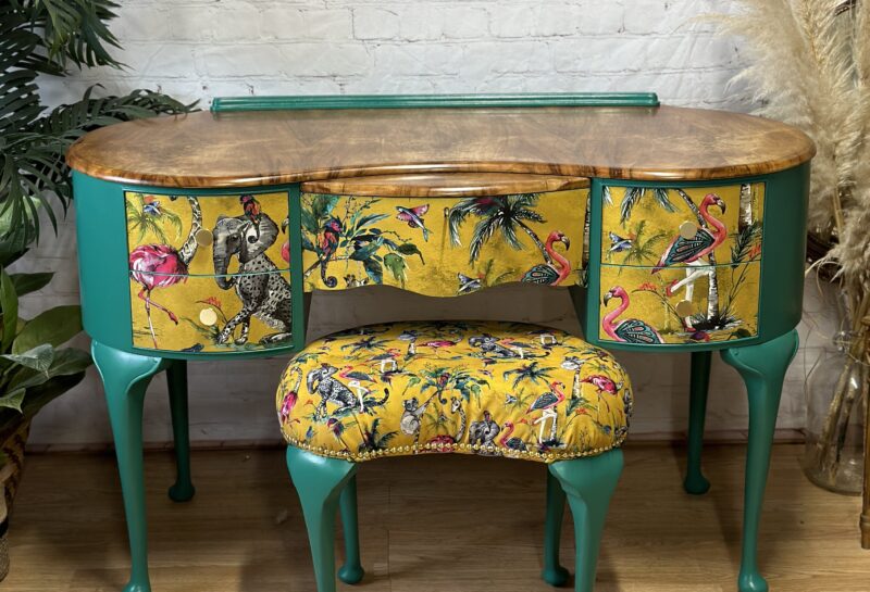 A vintage vanity table with vibrant green legs and sides, adorned with drawer fronts and a matching stool covered in yellow fabric with tropical bird and foliage patterns. The tabletop has a natural wood finish. Decorative elements include plants and a stylish mirror.