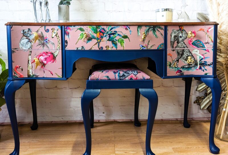 A vintage wooden desk with elegant curved legs and deep blue accents. The desk is adorned with colorful, vibrant wildlife-themed panels, featuring exotic birds, koalas, and flowers. A matching upholstered stool sits beneath it. Dried plants and candle on top.