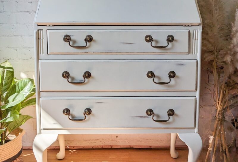 A vintage white secretary desk with an angled writing surface and three drawers featuring ornate brass handles. The desk is situated on a wooden floor, with a potted green plant to the left and tall, dried pampas grass in a vase to the right.