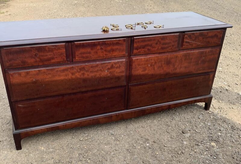 A vintage wooden dresser with a dark finish and eight drawers, placed outdoors on a gravel surface. The dresser's handles have been removed and are arranged on the top surface of the dresser. The handles appear to be metallic and are in multiple pieces.