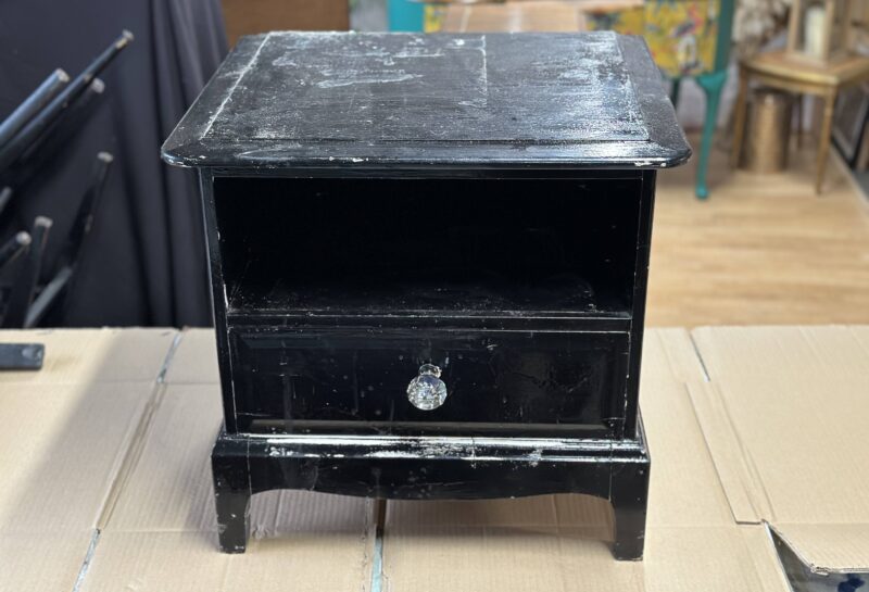 A weathered black wooden nightstand with a single drawer and an open shelf above it. The drawer has a round, crystal-like knob. The nightstand shows signs of wear and tear, including scratches and scuffed paint. It is placed on a cardboard surface.