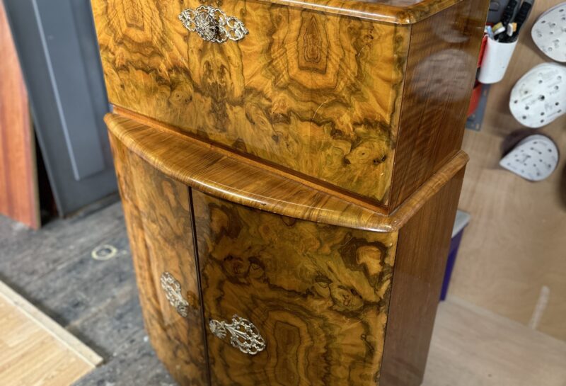 A wooden cabinet, featuring a highly polished finish with intricate patterns, stands in a workshop. The top section has two ornate metal handles, and the bottom section has two doors with similar handles. Various tools and supplies are visible in the background.