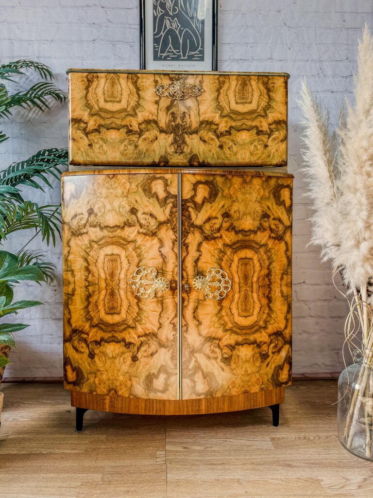 A vintage wooden cabinet with ornate patterns and metal handles stands against a white brick wall. The cabinet is flanked by potted plants on both sides, with tall pampas grass to the right and a leafy green plant to the left. A framed artwork hangs above the cabinet.