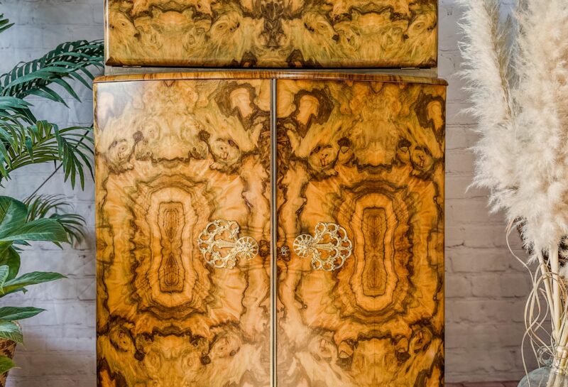 A vintage wooden cabinet with ornate patterns and metal handles stands against a white brick wall. The cabinet is flanked by potted plants on both sides, with tall pampas grass to the right and a leafy green plant to the left. A framed artwork hangs above the cabinet.