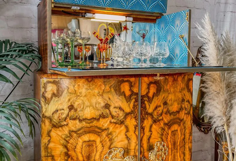 A vintage wooden cabinet with intricate grain patterns, open to reveal an assortment of glassware, including wine glasses and goblets. The interior is lined with blue and white geometric wallpaper. Plants and decorative elements are visible beside the cabinet.