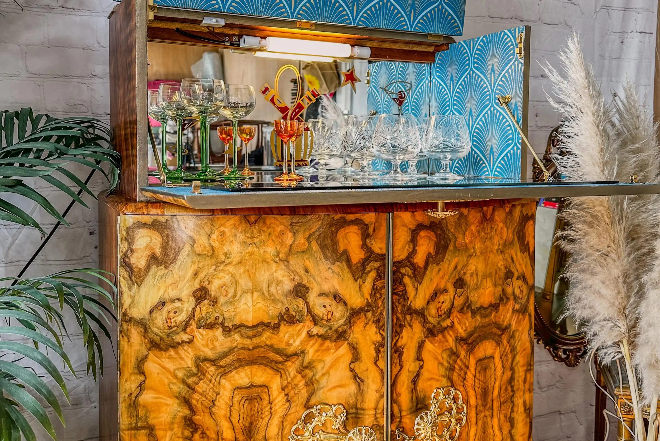 A vintage wooden cabinet with intricate grain patterns, open to reveal an assortment of glassware, including wine glasses and goblets. The interior is lined with blue and white geometric wallpaper. Plants and decorative elements are visible beside the cabinet.