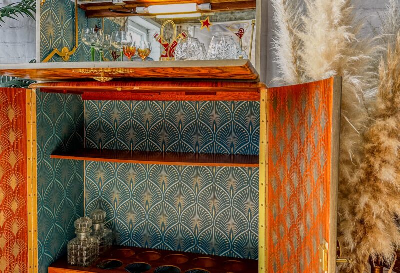 A vintage wooden cabinet adorned with colorful, geometric wallpaper is open to reveal decorative glassware and liquor bottles inside. Tall pampas grass stands to the right, and the background features a white brick wall with a framed picture above the cabinet.