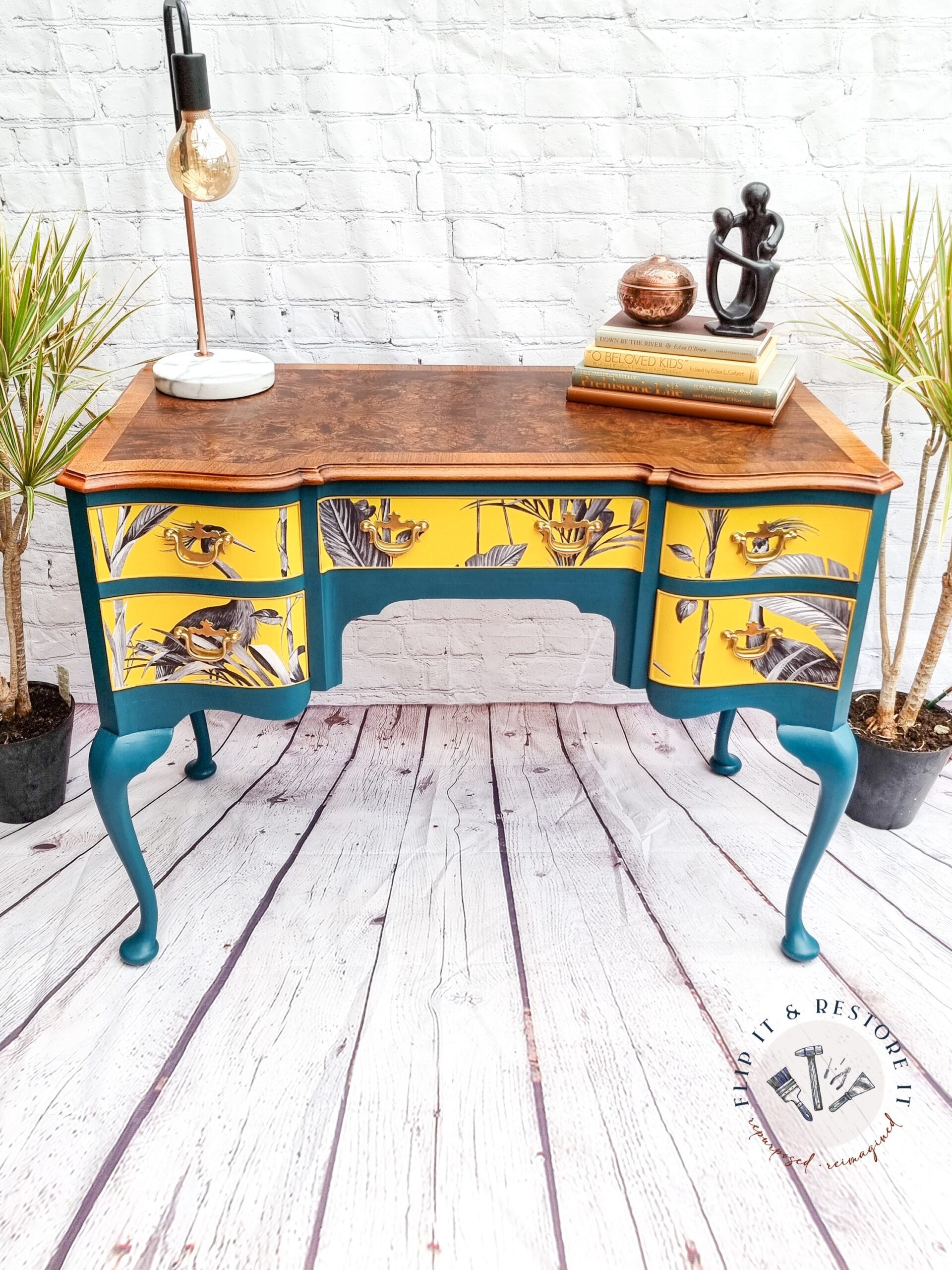 A vintage wood vanity table, refinished with teal legs and trim, yellow drawer faces featuring black and white floral designs. It has a polished wood top with a lamp, a stack of books, and a decorative sculpture. The table sits on a white wooden floor with two potted plants.