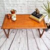 A G Plan Astro Mid Century Modern Vintage Teak Coffee Table from the 1960's with curved legs is placed against a white brick wall. On the table are books, a dog figurine, a potted plant, a white mug, a copper container, and a white pitcher with a wooden handle. The floor is wooden.