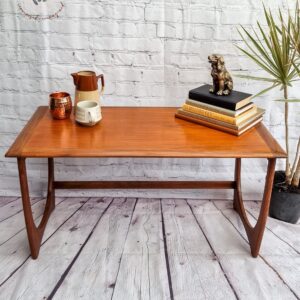 Against a white brick wall stands a G Plan Astro Mid Century Modern Vintage Teak Coffee Table from the 1960s, showcasing decorative angled legs. On the table sits a copper mug, a white mug, and a ceramic pitcher. A stack of books topped with a small bronze bear figurine and a potted plant add to the aesthetic. The table also features a logo in the top left corner.