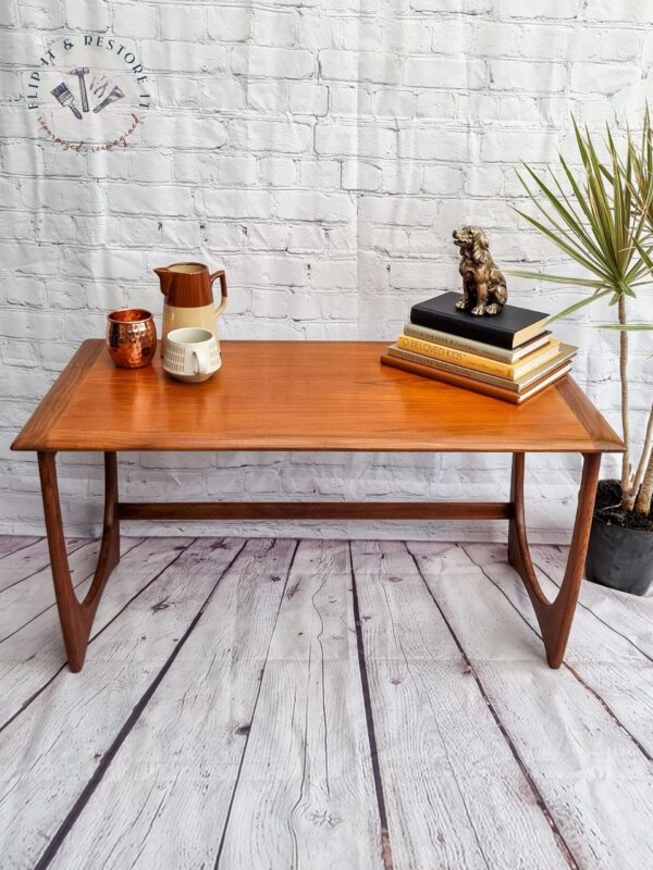 Against a white brick wall stands a G Plan Astro Mid Century Modern Vintage Teak Coffee Table from the 1960s, showcasing decorative angled legs. On the table sits a copper mug, a white mug, and a ceramic pitcher. A stack of books topped with a small bronze bear figurine and a potted plant add to the aesthetic. The table also features a logo in the top left corner.