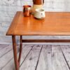 The G Plan Astro Mid Century Modern Vintage Teak Coffee Table from the 1960s, with its sleek, minimalist design, is set against a white brick wall. On top of the table are three mugs: a copper mug, a brown and tan ceramic pitcher, and a grayish-white ceramic mug. The corner of the table is visible, accentuating its elegant style.