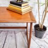 A G Plan Astro Mid Century Modern Vintage Teak Coffee Table from the 1960s holds a stack of books topped with a small gold cat sculpture, sitting next to a potted plant with long, slender leaves. The background features a white brick wall and the floor is made of wooden planks. A logo in the bottom right corner reads "BUILD IT RESTORE IT.