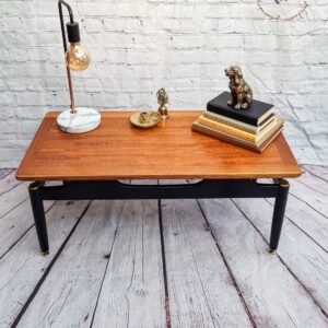 The G-Plan Librenza Mid Century Modern Black and Tola Vintage Coffee Table from the 1950s, adorned with a decorative lamp featuring a unique light bulb, gold accents, and a marble base, sits against a white brick wall with a circular logo. On the table are two gold sculptures, a small tray, and a stack of books.