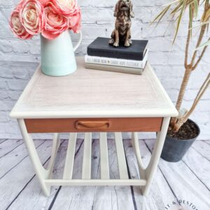 A Mid Century Beautility Drinks Cabinet from the 1960s, made of walnut and designed for use as a vintage cocktail bar, features a vase containing pink and white flowers, two stacked books, and a dog statuette. The cabinet sits on a wooden floor with a brick wall background and is accompanied by a potted plant on the right.