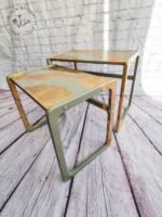 Two G-Plan Style Mid Century Nest of Two Side Tables, featuring worn, rustic wooden tops and green metal legs, stand on a pale wooden floor against a white brick wall. The tables have a weathered appearance with patches of exposed wood, giving them a distressed, shabby-chic look.