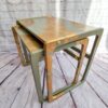 Two G-Plan Style Mid Century Nest of Two Side Tables with a rustic, distressed paint finish in green and gold. The tables are placed on a wooden floor against a white brick wall background. The logo “Rye the Renovator" is visible in the upper left corner of the image.