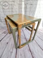 Two G-Plan Style Mid Century Nest of Two Side Tables with a rustic, distressed paint finish in green and gold. The tables are placed on a wooden floor against a white brick wall background. The logo “Rye the Renovator" is visible in the upper left corner of the image.