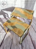 A pair of G-Plan Style Mid Century Nest of Two Side Tables featuring worn green and gold paint. The tables are positioned on a wooden floor against a brick wall, with their paint chipped and peeling to reveal a rustic, distressed look. The shadow cast by the table legs creates interesting patterns.