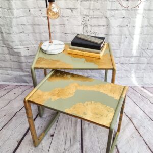 Two **G-Plan Style Mid Century Nest of Two Side Tables** with a green and gold abstract design are placed on a wooden floor against a white brick wall. The top table holds a lamp with an exposed bulb, marble base, and a stack of books. The wall displays a "Flip It & Restore It" logo.