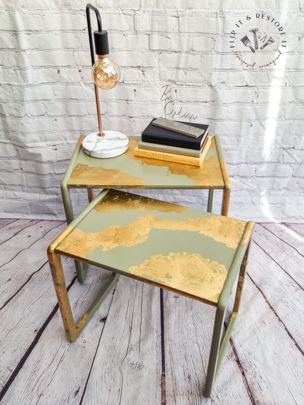 Two **G-Plan Style Mid Century Nest of Two Side Tables** with a green and gold abstract design are placed on a wooden floor against a white brick wall. The top table holds a lamp with an exposed bulb, marble base, and a stack of books. The wall displays a "Flip It & Restore It" logo.