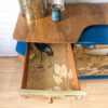 A Mid Century Beautility Drinks Cabinet from the 1960s stands against a white brick wall, adorned with a brass vase and ceramic figure on top. One drawer is open, revealing a floral and bird pattern inside. The cabinet has a blue accent and sits on a laminate wood floor.