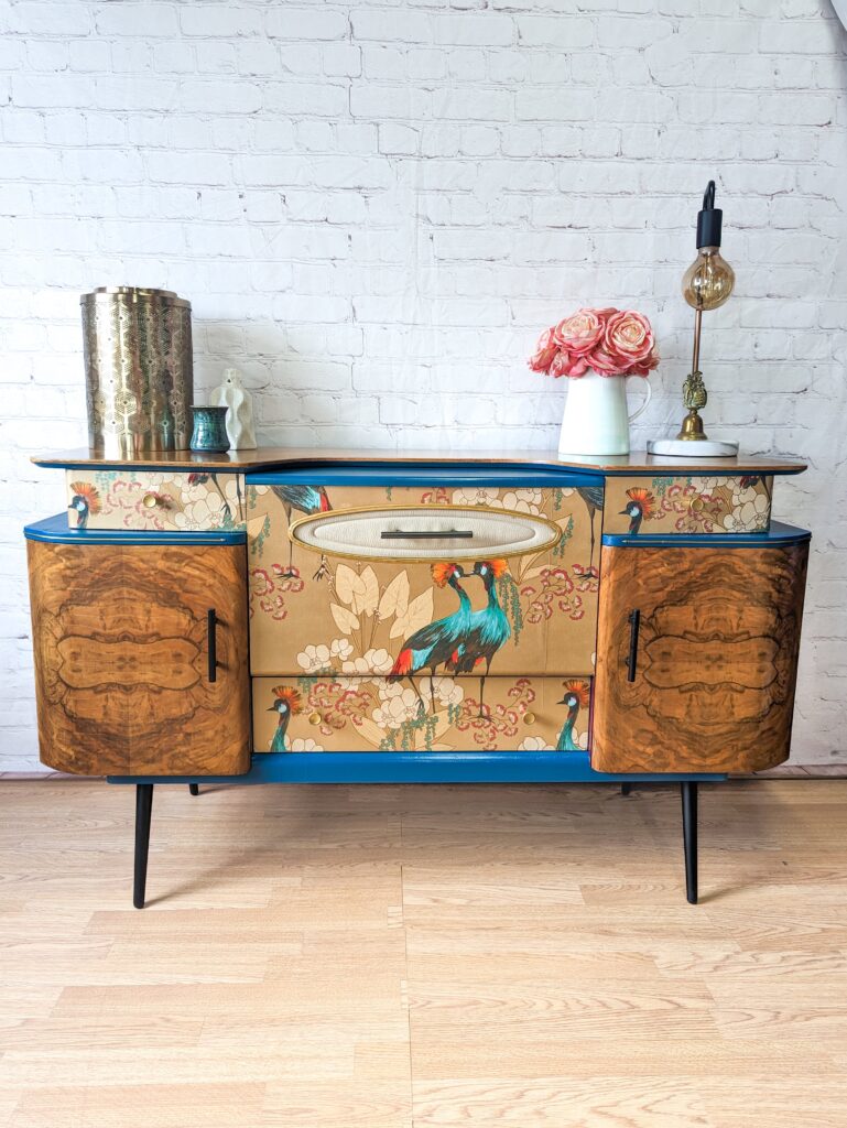 The Mid Century Beautility Drinks Cabinet, a vintage 1960's walnut cocktail bar, showcases peacock artwork on its doors and drawers. It features teal accents and slender black legs. Atop the cabinet sit a gold cylindrical vase, a small white vase with pink flowers, and a lamp with an exposed bulb.