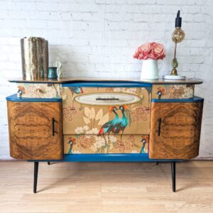 The Mid Century Beautility Drinks Cabinet, a vintage 1960's walnut cocktail bar, showcases peacock artwork on its doors and drawers. It features teal accents and slender black legs. Atop the cabinet sit a gold cylindrical vase, a small white vase with pink flowers, and a lamp with an exposed bulb.