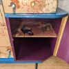 A close-up view of the Mid Century Beautility Drinks Cabinet, Vintage 1960's Walnut Cocktail Bar. The cabinet exterior boasts a vibrant patterned design with birds and flowers. One section is open, showcasing a purple-painted interior and a shelf adorned with similar bird-themed artwork. The wooden floor beneath adds to the overall aesthetic appeal.