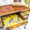 The Queen Anne Painted Burr Walnut Dressing Table/Vanity/Desk/Sideboard is adorned with books and a sculpture. One drawer is open, revealing a vibrant yellow interior with a black and white bird and plant design. The desk features teal accents and brass handles, set against a white brick wall and sumac flooring.