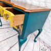 Against a white brick wall, the Queen Anne Painted Burr Walnut Dressing Table/Vanity/Desk/Sideboard showcases its teal-painted legs and sides with a natural wood top. One drawer is partially open, revealing a vibrant yellow interior adorned with decorative patterns and featuring a vintage-style brass handle. An artist's emblem rests on the floor nearby.