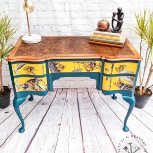 A small, vintage Queen Anne Painted Burr Walnut desk with teal legs and three drawers, each showcasing vibrant yellow floral and bird designs. The top is made of dark-finished wood. The desk is styled with a lamp, two books, a decorative sculpture, and a potted plant.