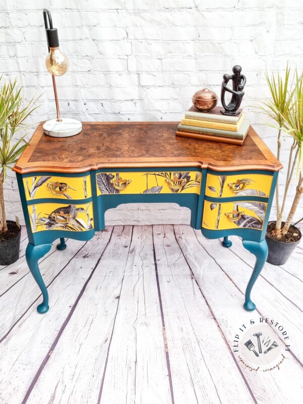 A small, vintage Queen Anne Painted Burr Walnut desk with teal legs and three drawers, each showcasing vibrant yellow floral and bird designs. The top is made of dark-finished wood. The desk is styled with a lamp, two books, a decorative sculpture, and a potted plant.