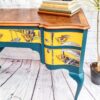 A vibrantly painted Queen Anne Painted Burr Walnut Dressing Table/Vanity/Desk/Sideboard featuring teal and yellow drawers adorned with floral and bird designs. The surface holds a stack of books, a decorative plant, and a small sculpture. The scene is set against a white brick wall and wooden floor.