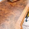 A Queen Anne Painted Burr Walnut dressing table, vanity, desk, or sideboard with a richly textured, polished surface. Books are neatly stacked on one side, including titles like "Prehistoric Life" and "Loved Kids." The edge of the desk features intricate details, and a logo in the corner reads "Flip It & Restore It.