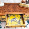 A Queen Anne Painted Burr Walnut Dressing Table/Vanity/Desk/Sideboard against a white brick wall. The desk features intricate grain patterns and a drawer that is open, revealing a vibrant yellow interior with a black and white bird and reed design. Books, including one titled "Beloved Kids," are stacked on top.