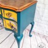 A Queen Anne Painted Burr Walnut Dressing Table/Vanity/Desk/Sideboard featuring teal-painted cabriole legs and sides, with two drawers that have yellow fronts adorned with feather designs. The tabletop showcases a rich wood grain finish. A small logo in the bottom right corner reads "Flip It & Restore It," against a white brick background.