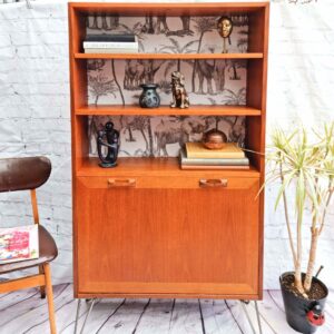 A G-Plan Sierra Teak Bookcase Drinks Cabinet with hairpin legs showcases two shelves adorned with decorative items, including a gold mask, a ceramic owl, and small animal figurines. The back panel is enhanced with black-and-white animal-themed wallpaper. Below the shelves lies an enclosed storage area. The scene also includes a chair, a book, and a potted plant.