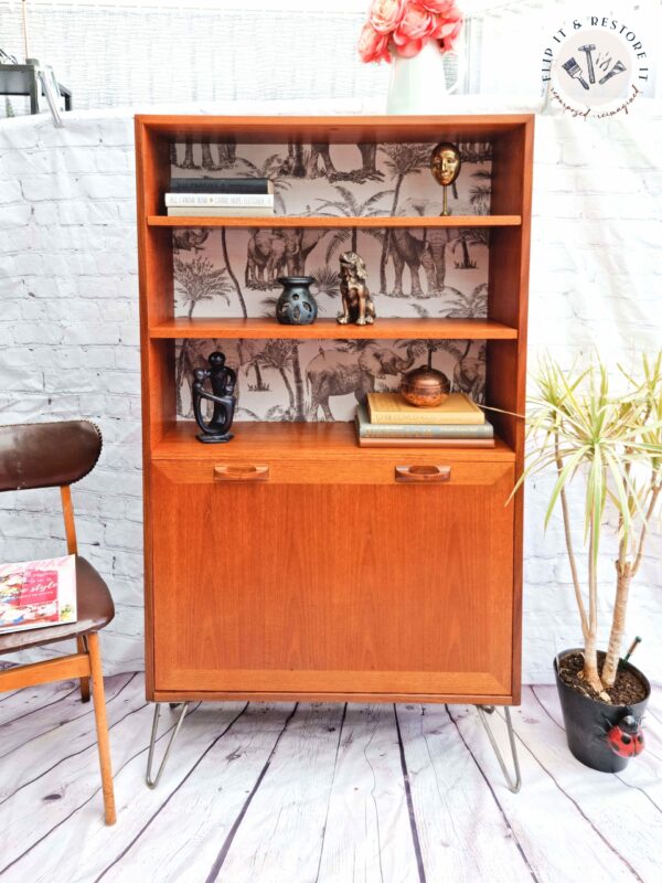 A G-Plan Sierra Teak Bookcase Drinks Cabinet with hairpin legs showcases two shelves adorned with decorative items, including a gold mask, a ceramic owl, and small animal figurines. The back panel is enhanced with black-and-white animal-themed wallpaper. Below the shelves lies an enclosed storage area. The scene also includes a chair, a book, and a potted plant.