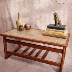 A Vintage Mid Century Solid Teak Tiled Two-Tier Coffee Table from the 1970’s adorned with decorative items: a small statue of a dog perched on stacked books, a brown glass vase, a green candle, and a tall vase with a woven texture. The table is set on a brown carpet against a beige backdrop and logo.