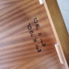 A close-up of the interior of a wooden drawer from the Mid Century Teak Nest of Tables - Coffee Table and Two Side Tables, showcasing its unique wood grain pattern. The text "EN I K-79 CL" is stamped in black ink on the inner surface. The drawer features wooden guides and is partially opened, revealing its construction details.