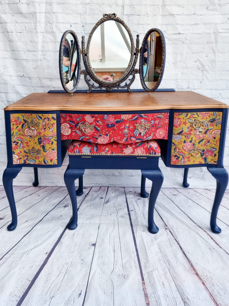 A vintage vanity with navy blue legs and a wooden tabletop stands against a white brick wall. The drawers feature vibrant red and gold floral patterns. An ornate, triple-panel mirror sits on top, with reflections visible in the mirror.