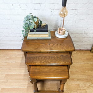 The G-Plan Astro Round Coffee Table Mid-Century Vintage is adorned with a stack of books, a small potted plant, and a decorative giraffe figurine. An Edison bulb lamp, a ceramic candle, and a leafy vine plant complete the arrangement. The background features a white brick wall and hardwood flooring.