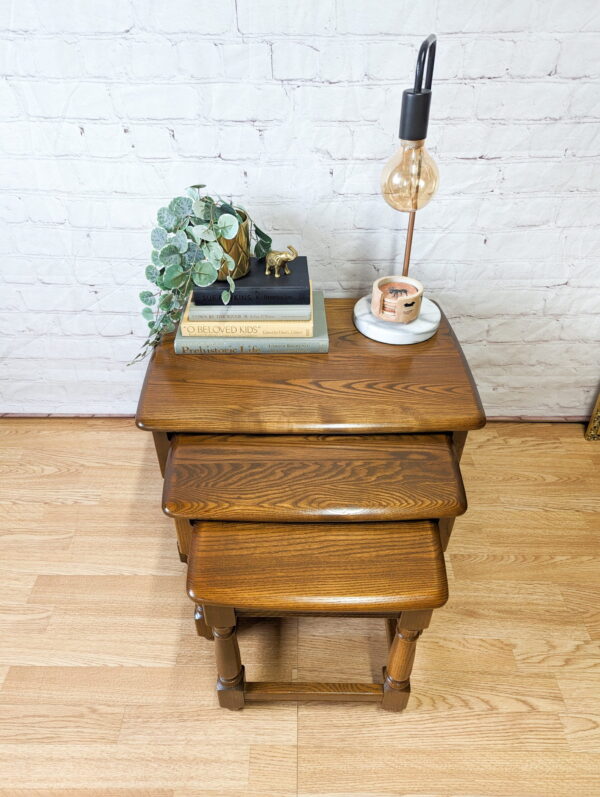 The G-Plan Astro Round Coffee Table Mid-Century Vintage is adorned with a stack of books, a small potted plant, and a decorative giraffe figurine. An Edison bulb lamp, a ceramic candle, and a leafy vine plant complete the arrangement. The background features a white brick wall and hardwood flooring.