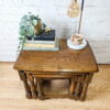 The G-Plan Astro Round Coffee Table Mid-Century Vintage holds a stack of books, a potted plant, a small figurine, and a lamp. Next to the books is a circular tray with a candle. The background features a white brick wall and a wooden floor.