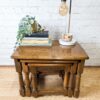 A G-Plan Astro Round Coffee Table Mid-Century Vintage set against a white brick wall holds a small collection of items, including a green potted plant, stacked books, a small decorative elephant, and a modern desk lamp with an exposed bulb. The larger table has neatly tucked the smaller table beneath it.