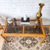 An Ercol Saville Ash Long Coffee Table with Smoked Glass Mid Century Vintage sits on an ornate rug. On the table are a candle, a small statue, books, a white geometric vase, and a tall brass vase. Behind the table is a white brick wall.