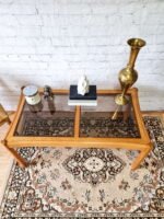 An Ercol Saville Ash Long Coffee Table with Smoked Glass Mid Century Vintage sits on an ornate rug. On the table are a candle, a small statue, books, a white geometric vase, and a tall brass vase. Behind the table is a white brick wall.