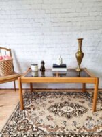 A room with a white brick wall features the Ercol Saville Ash Long Coffee Table with Smoked Glass Mid Century Vintage. Items on the table include a candle, a small sculpture, a stack of books, and a golden vase. A wicker chair with a cushion and a patterned rug are also visible.